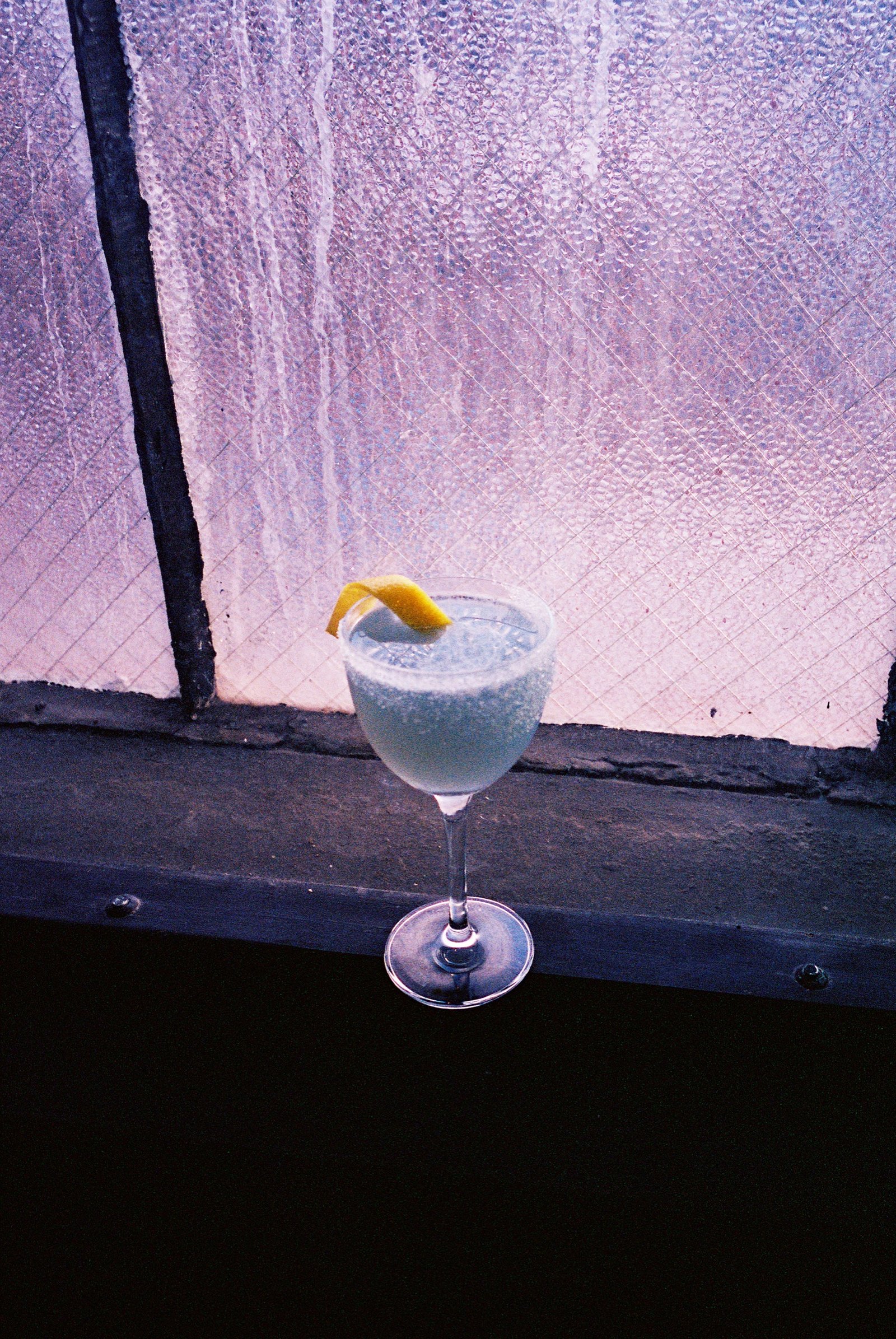Lemon-twist cocktail on a sill against violet light through textured glass